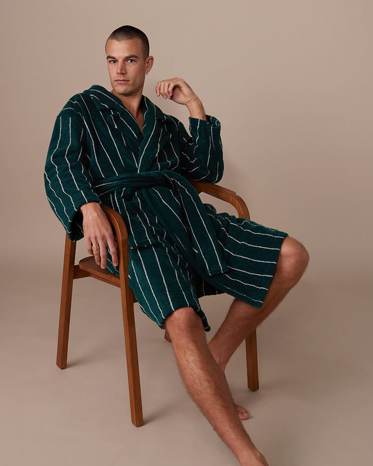 Man wearing a green Hommey robe sitting on a wooden chair against a beige background