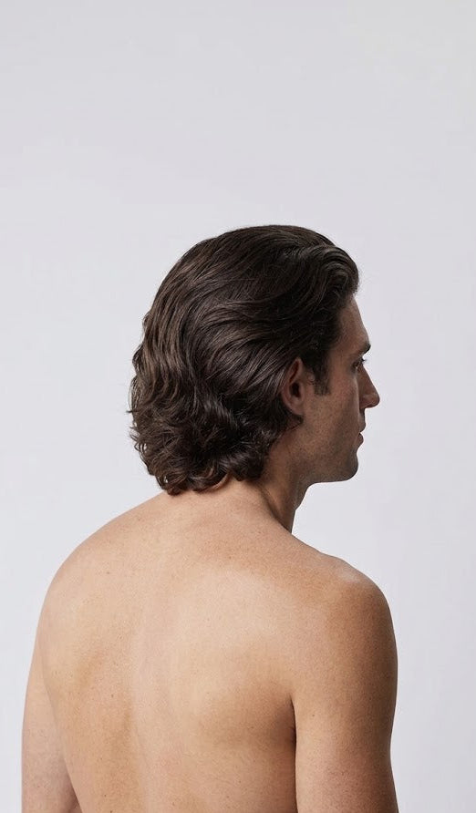 A man with wavy dark brown hair, shown from a side-back profile against a light gray studio background.
